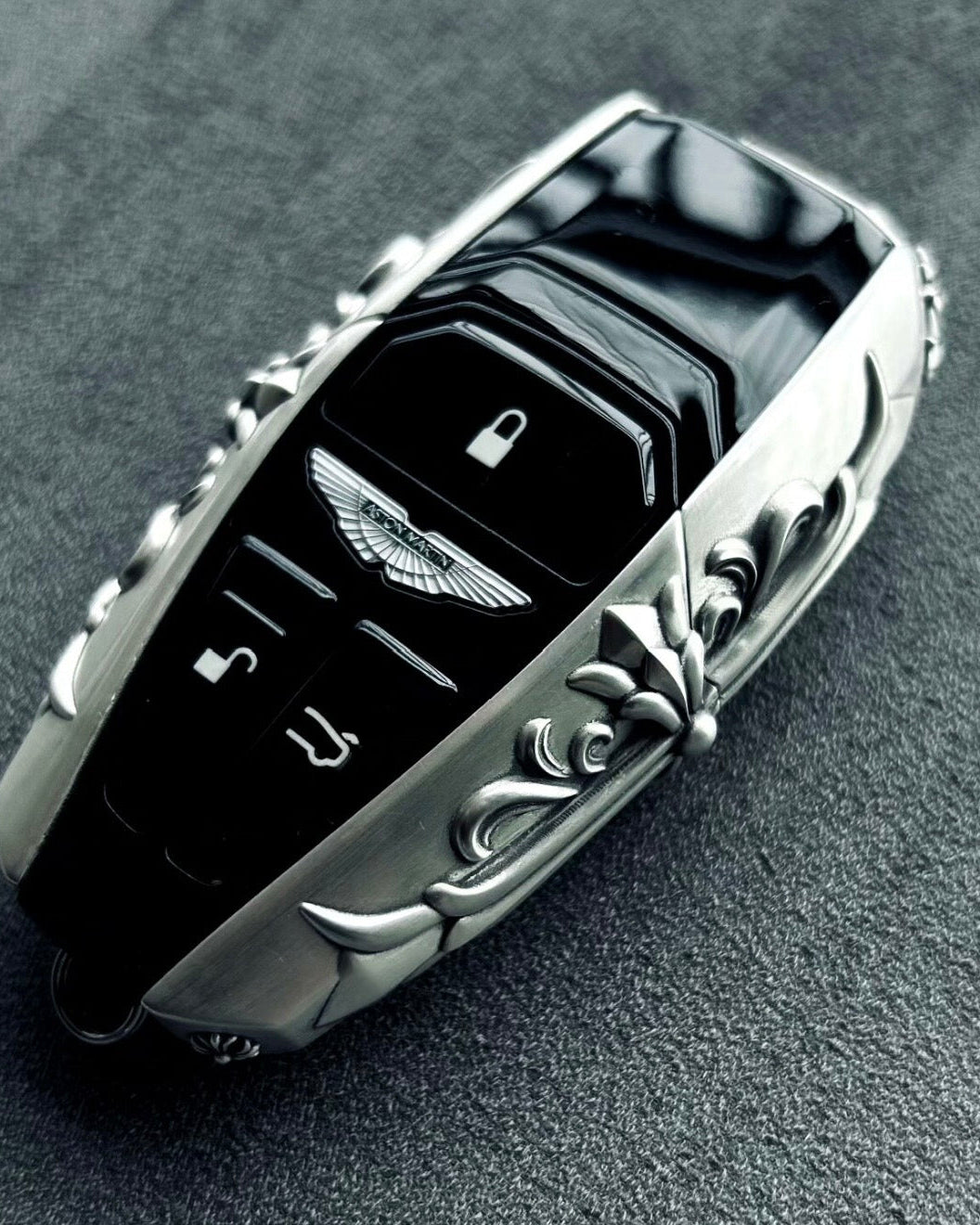 Aston Martin handcrafted sterling silver key case 45-degree angle view showing iconic winged emblem, three-button layout and ornate engraved solid silver frame with refined detailing