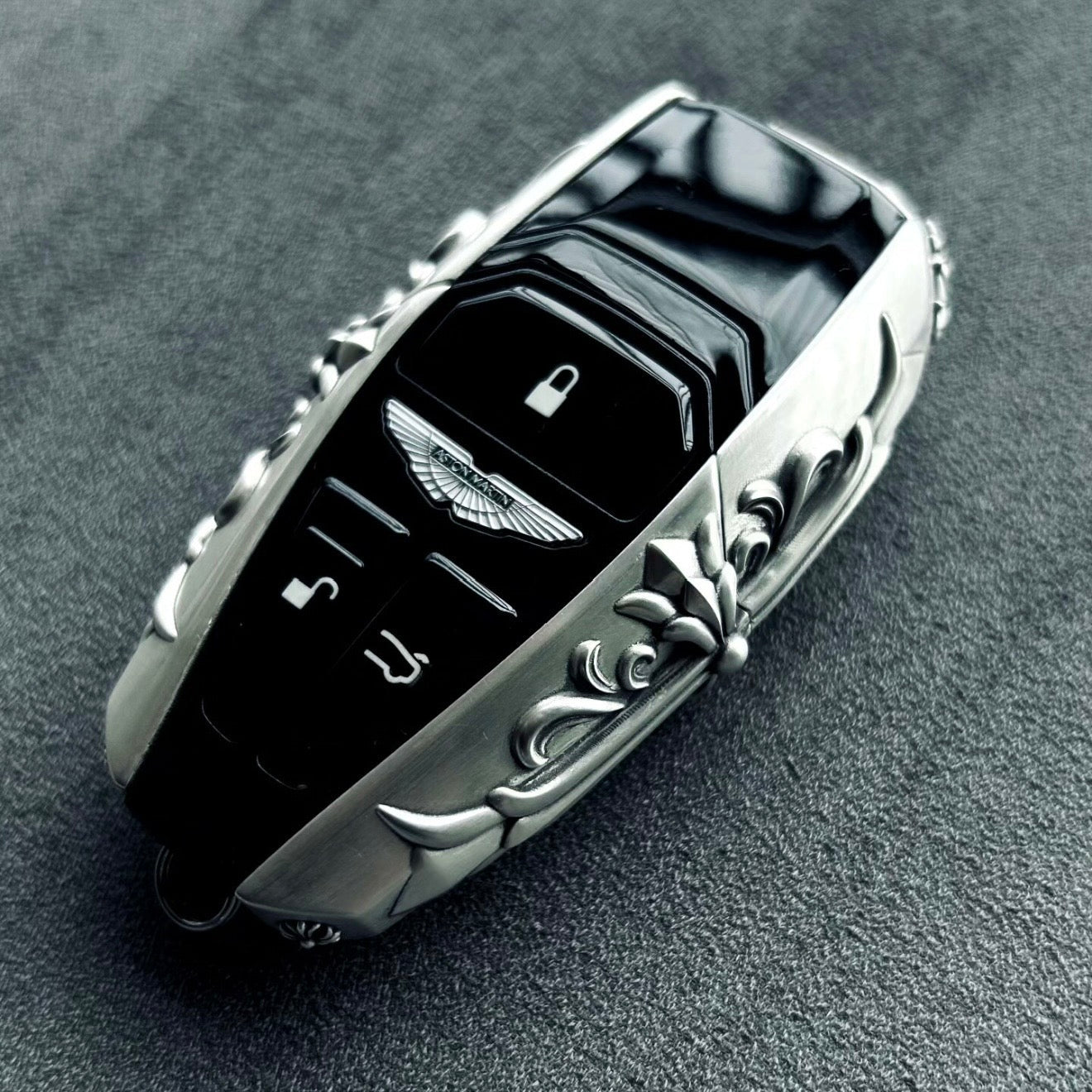 Aston Martin handcrafted sterling silver key case 45-degree angle view showing iconic winged emblem, three-button layout and ornate engraved solid silver frame with refined detailing