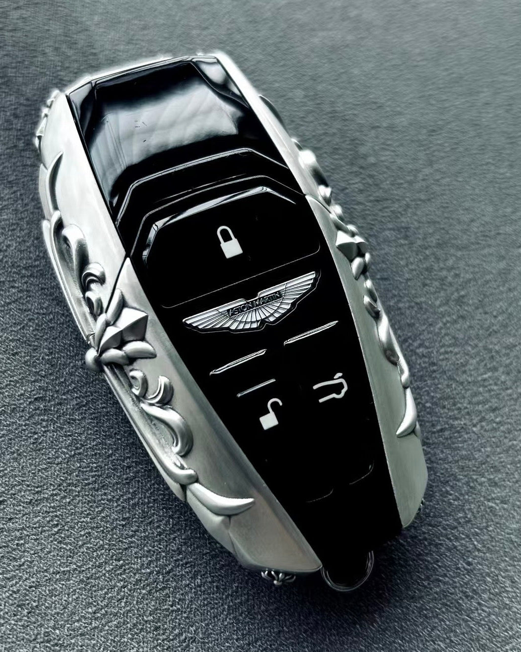 Aston Martin handcrafted sterling silver key case front view featuring iconic Aston Martin winged emblem, lock/unlock/trunk buttons and symmetrical engraved precious metal frame
