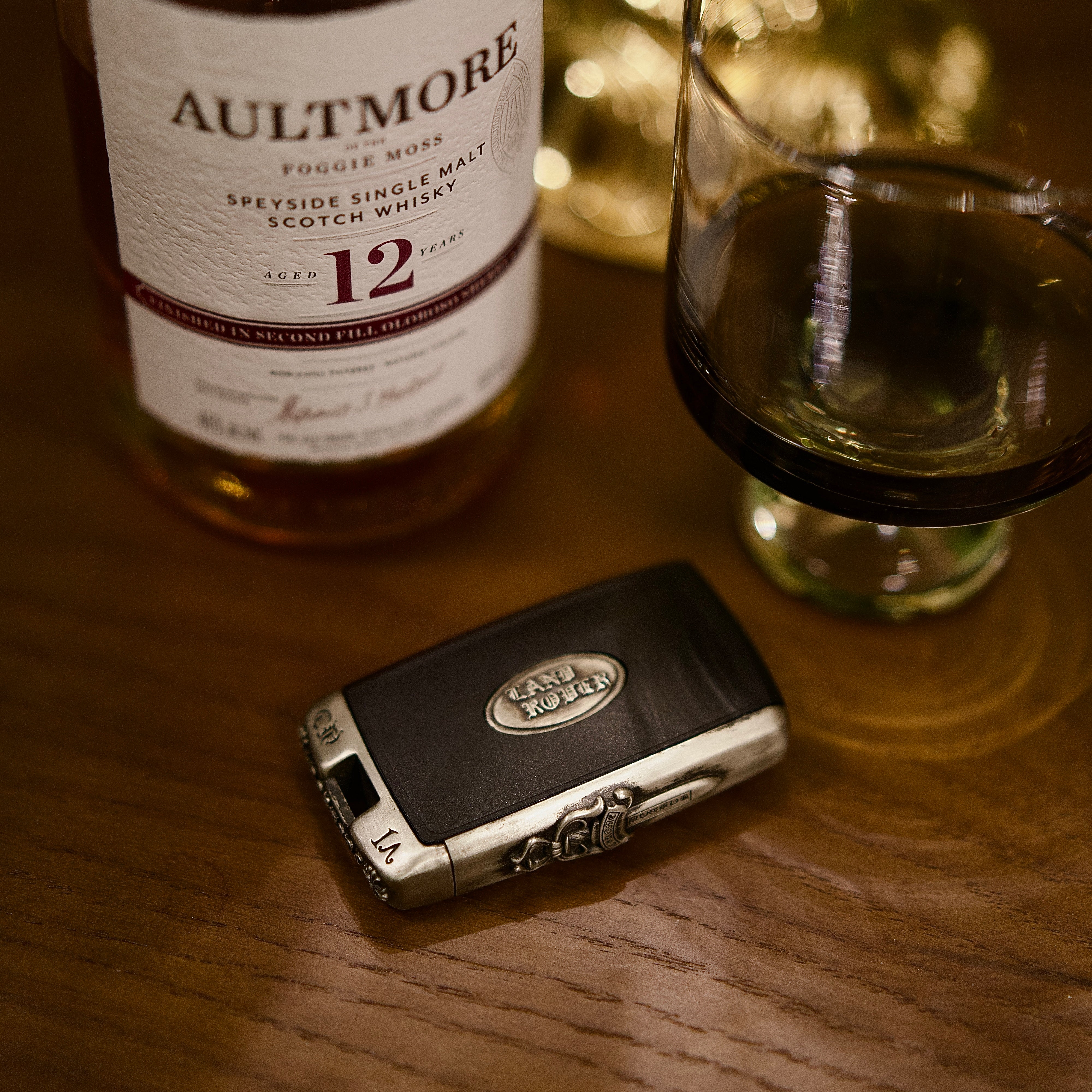 Luxury lifestyle shot: Land Rover key case displayed with AULTMORS 12 whiskey bottle and glass on wooden table, elegant ambiance showcasing refined taste