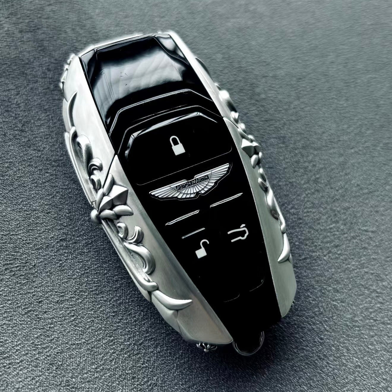 Aston Martin handcrafted sterling silver key case front view featuring iconic Aston Martin winged emblem, lock/unlock/trunk buttons and symmetrical engraved precious metal frame