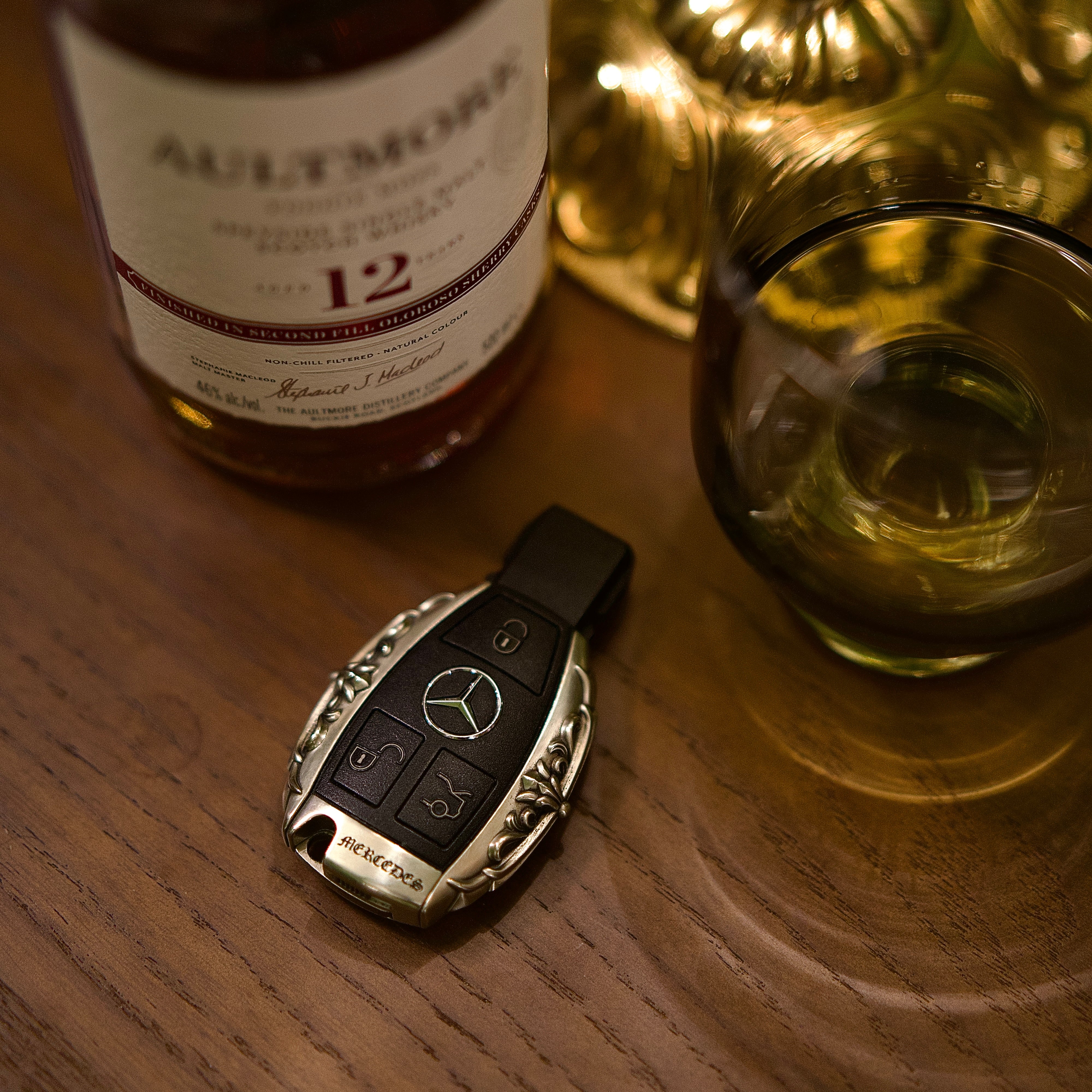Handmade silver key case for Mercedes flat lay on wooden table with whiskey glass warm ambient lighting showcasing masculine heft and understated luxury