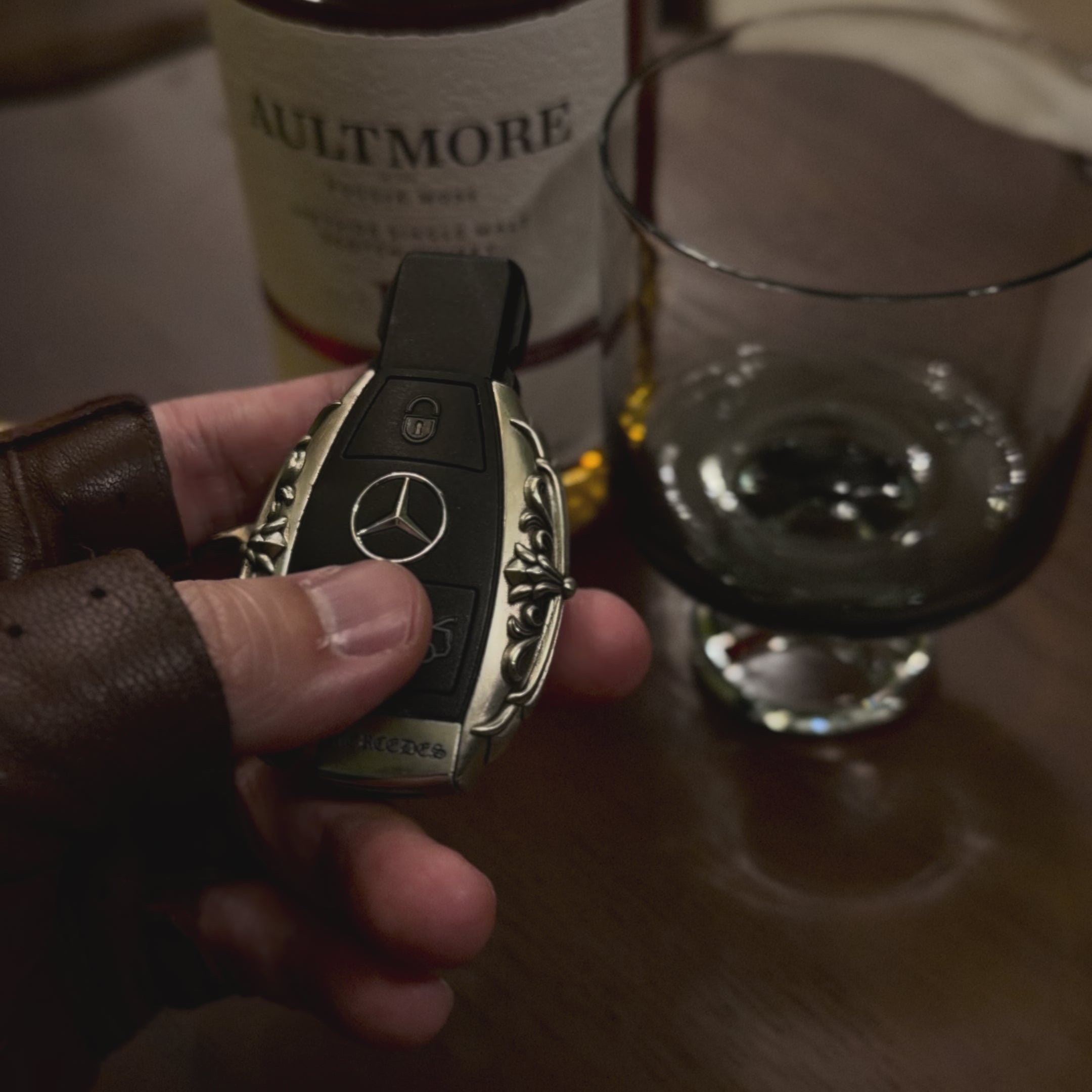 Substantial silver key case for Mercedes owner handheld perspective next to whiskey bottle showcasing considerable presence in daily carry and refined taste