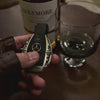 Substantial silver key case for Mercedes owner handheld perspective next to whiskey bottle showcasing considerable presence in daily carry and refined taste