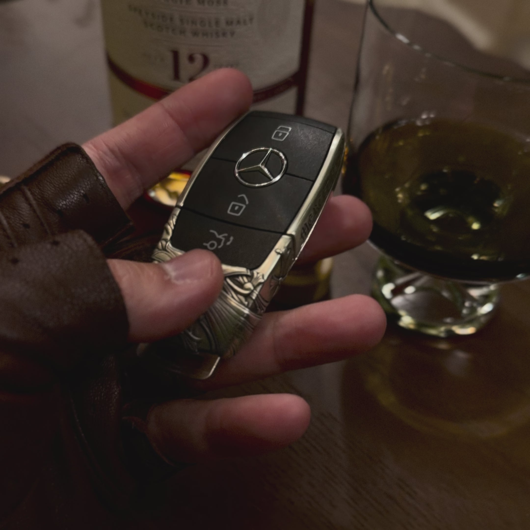 Substantial silver key case for Mercedes owner handheld perspective next to whiskey glass showcasing considerable presence in daily carry